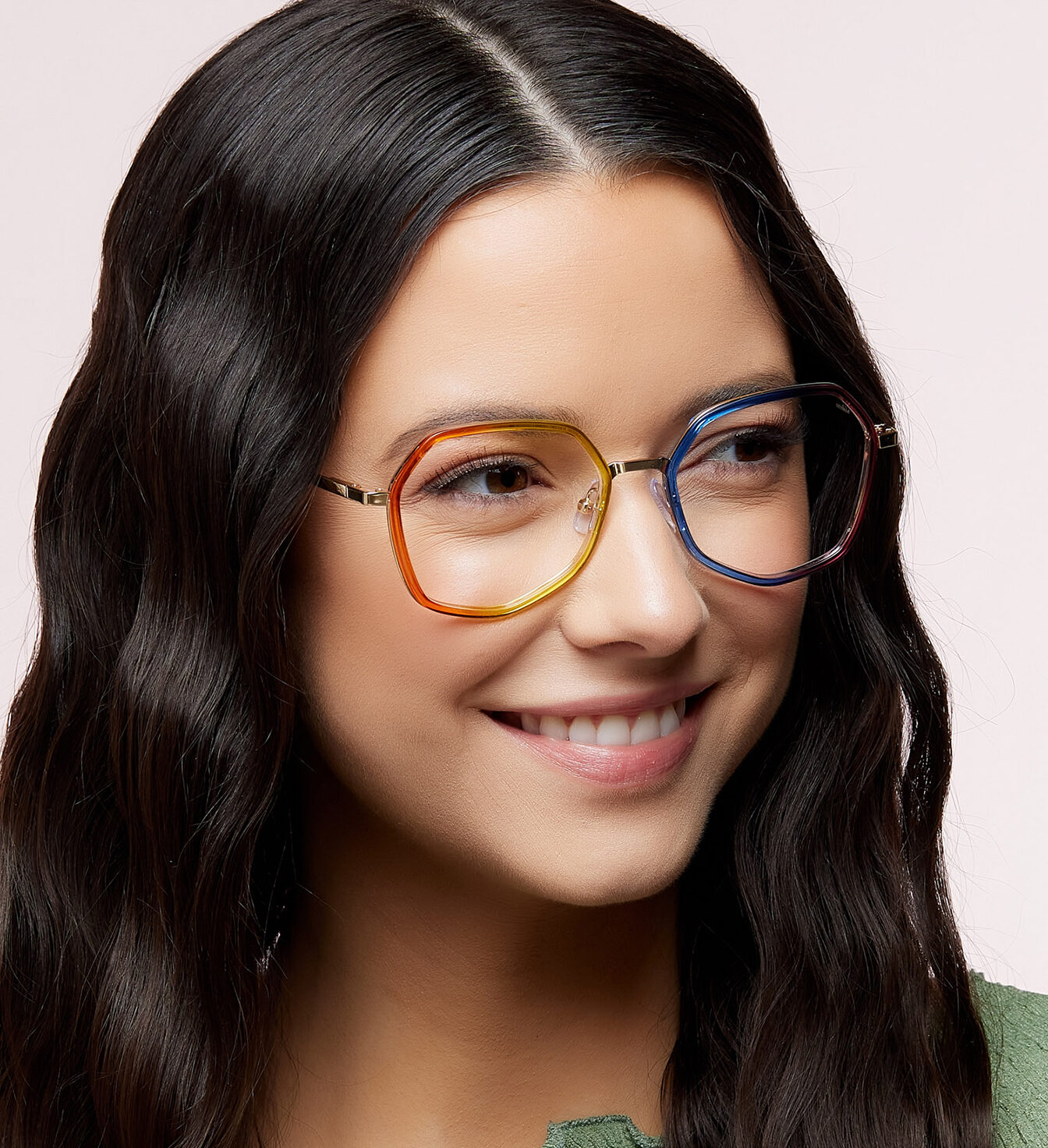 A smiling woman with long wavy dark hair wears geometric eyeglasses with rainbow-coloured frames and a green top, looking slightly to the side against a plain background.