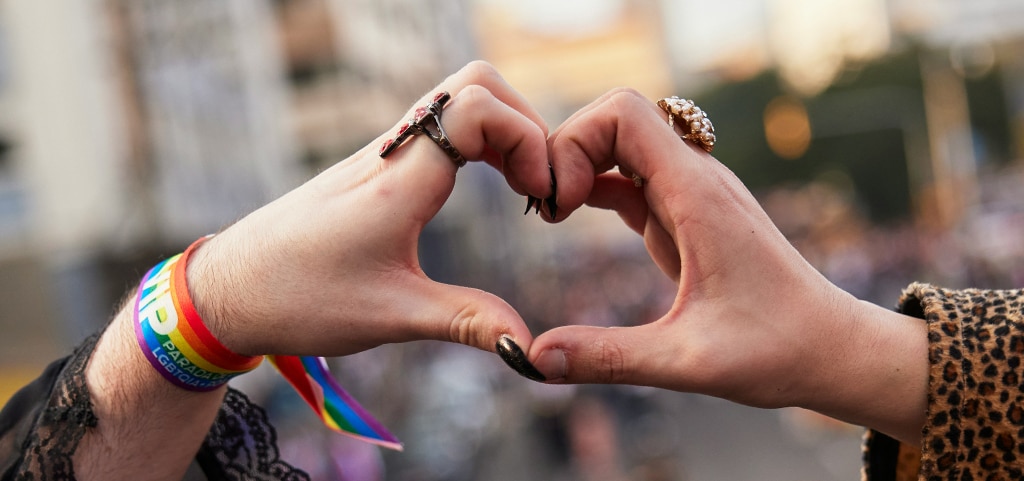 Two hands join to form a heart shape, one adorned with a black lace sleeve, a rainbow wristband, a ring and long black-painted nails; the other styled with a leopard-print sleeve and a ring.