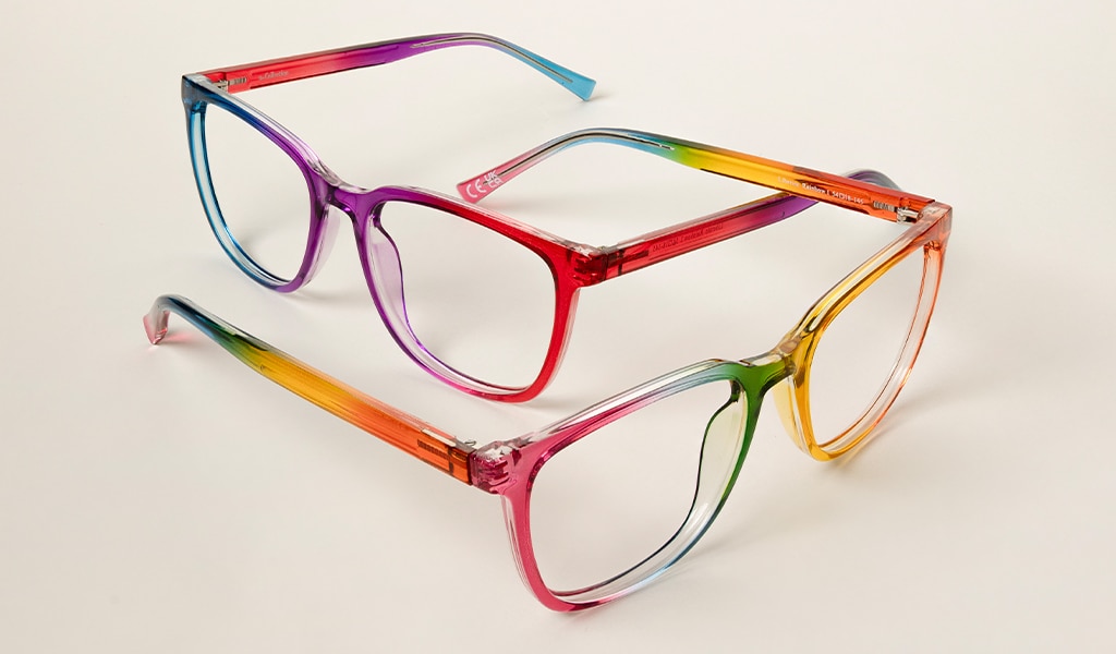Two rainbow-coloured square acetate glasses lie intertwined on a cream backdrop, each pair open and angled slightly apart.