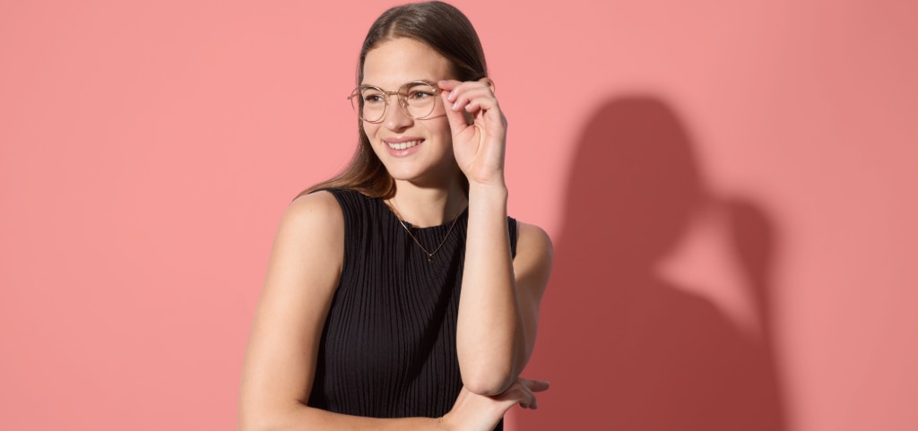 A portrait shot of a young lady with long. straight brown hair, standing in front of salmon-pink background. She is wearing a black pleated vest top and silver round metal glasses, with her left hand gently resting on the frames.