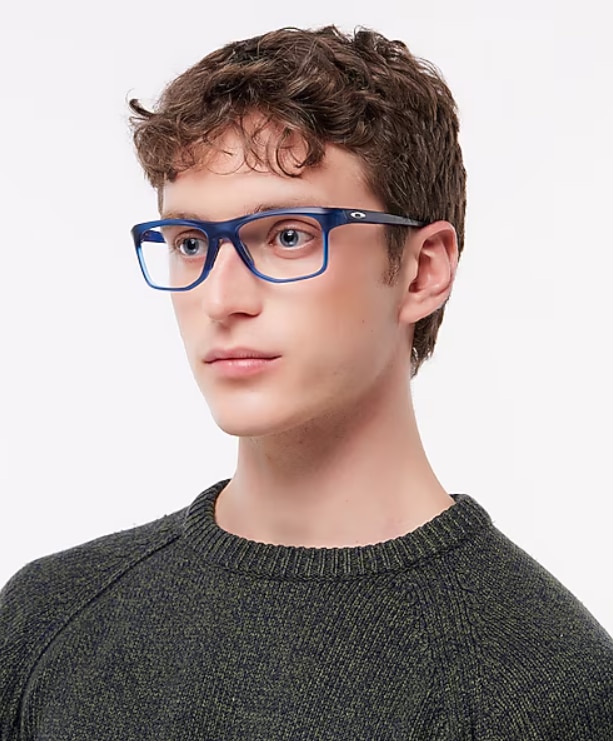 A young man with short curly brown hair wears blue rectangular Oakley eyeglasses and a dark sweater, looking slightly to the side with a neutral expression against a plain background.