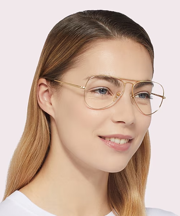 A woman with straight light-brown hair wears thin gold metal Ray-Ban Aviator eyeglasses with a white t-shirt, smiling gently and looking slightly to the side against a plain background.