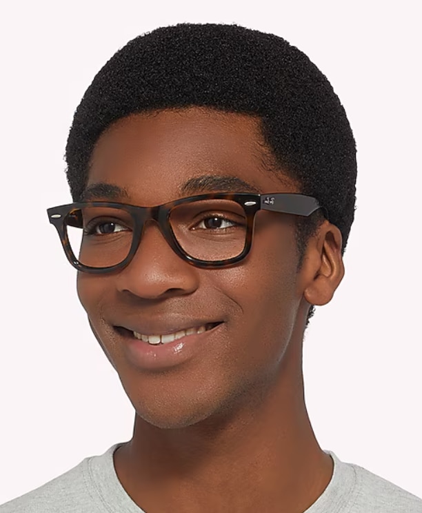 A smiling man with a short black afro wears dark Havana Ray-Ban Wayfarer glasses and a grey t-shirt, looking slightly to the side against a plain background.