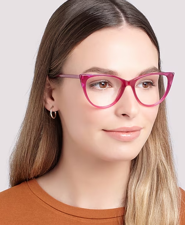 A woman with long light-brown hair wears pink cat-eye eyeglasses, small hoop earrings, and an orange t-shirt, looking slightly to the side with a soft, neutral expression against a plain background.