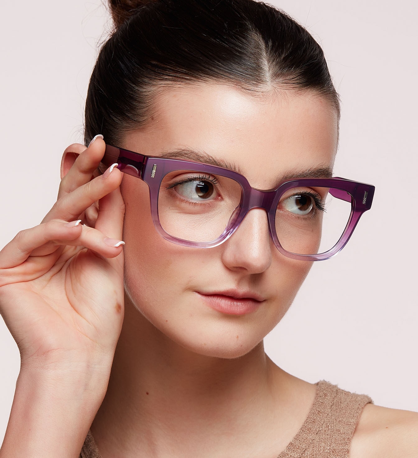 A woman with dark hair tied neatly back in a bun wears bold purple eyeglasses with a light-brown vest top, gently touching the frame while looking off to the side against a plain background.