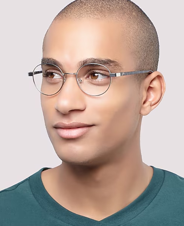 A man with a shaved head wears thin, round metal eyeglasses and a green V-neck t-shirt, looking slightly to the side with a calm expression against a plain background.