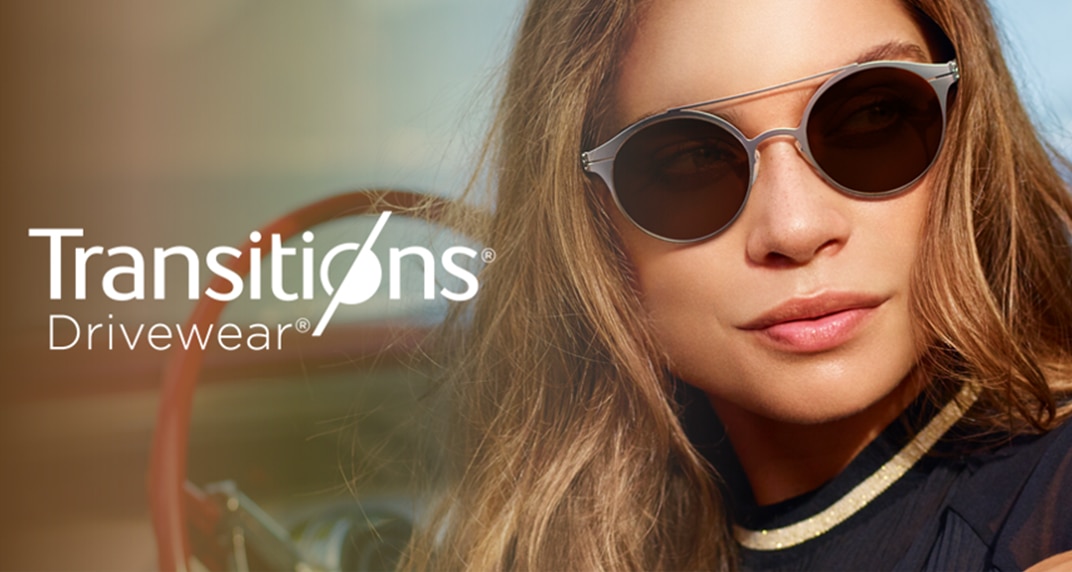 An image displaying the Transitions® Drivewear® logo in white over a background which shows a young lady behind the steering wheel of a car. She is facing away, toward the camera and has long brown hair. The lady is wearing a black top and round silver sunglasses with a double bridge design.