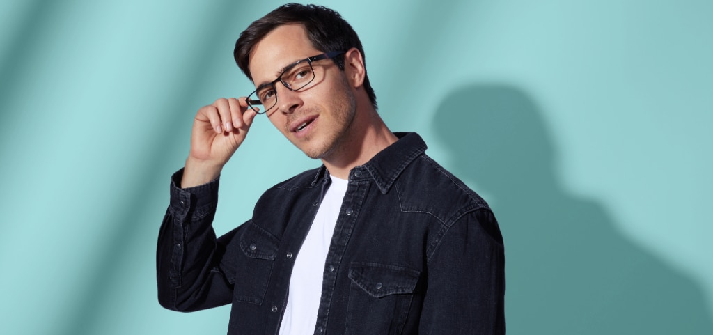 A man with short dark hair stands against a light turquoise background, wearing a black denim shirt layered over a white T-shirt. He sports black metal rectangular glasses and gently touches the frame with his right hand.