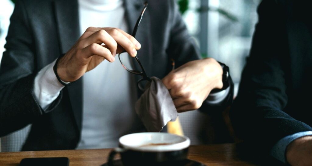 A close-up of someone cleaning their glasses with a cloth, dressed in a white turtleneck sweater under a grey blazer. Another person’s arm is partially visible on the left and a cup of coffee sits in front of them.