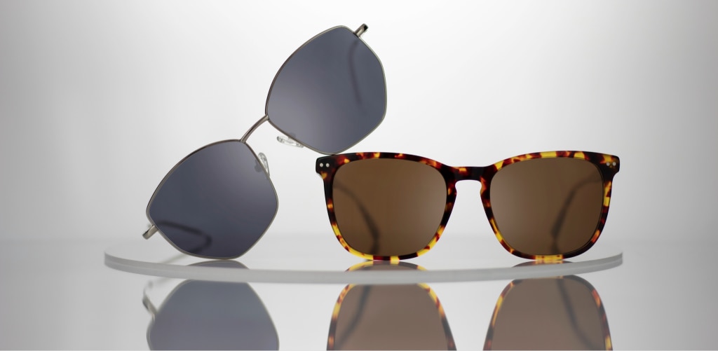 Two pairs of sunglasses are displayed on a curved clear stand against a light grey background. On the left, the GD Eliza geometric sunglasses feature grey-tinted lenses and silver metal arms, leaning diagonally. On the right, the GD Elsie sunglasses have square tortoiseshell frames with brown lenses and stand upright. Both pairs are reflected on the glossy surface below.