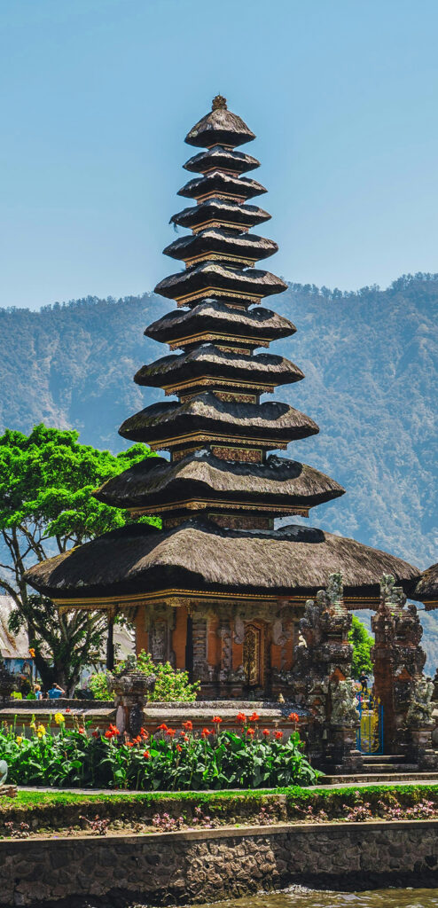 A photo of a temple in Bali. It has a multi-tiered, thatched-roof and is located on the waterfront. There are mountains visible among mist in the background.