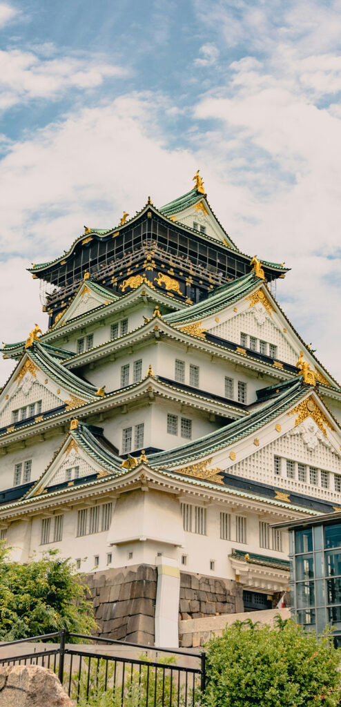 A photo of Osaka Castle's main tower, a five-story building with emerald-coloured roofs, ornate detailing and gold trim. Green foliage surrounds the site.