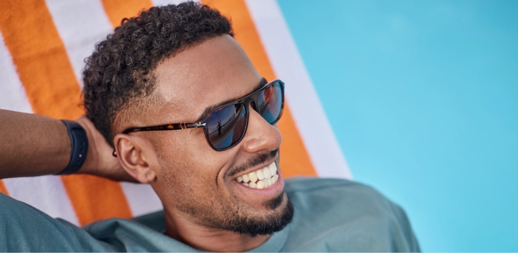 A smiling man wearing Persol PO3359V dark tortoiseshell square polarised sunglasses relaxes on a white-and-orange striped beach chair beside bright blue water on a sunny day.