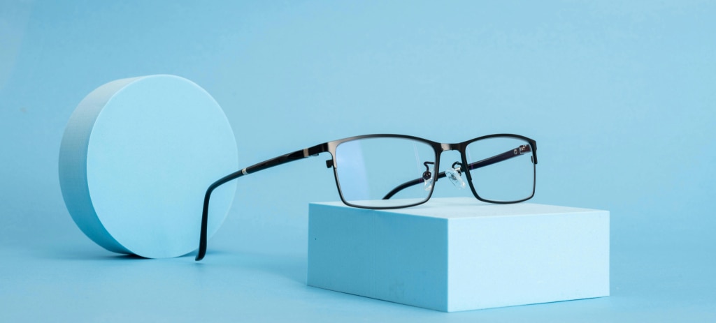 A pair of black metal rectangular glasses on a light-blue background. The glasses are balanced on a light-blue block and there is a cylinder behind them. 
