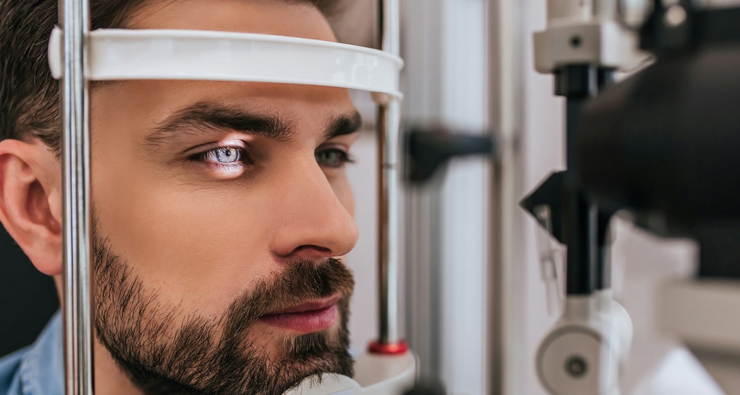 “A close-up of a young man undergoing an eye exam, with his chin resting on a slit lamp. He has dark brown hair, blue eyes, and a neatly trimmed beard. A bright light is focused on his right eye.