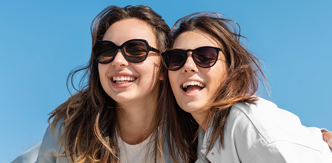 A close-up shot of two adult sisters. smiling brightly and embracing one and other. They are standing beneath a bright blue sky. Both have light brown hair and are wearing light-blue denim shirts paired with black sunglasses.
