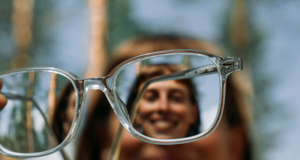 A close-up of transparent acetate glasses, with a woman's smiling face softly blurred but visible through the lenses. The scene is bathed in natural light, suggesting a bright outdoor setting.