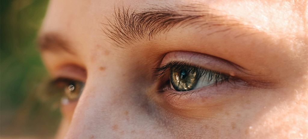 A close-up of the side of a person's face, highlighting their green-blue eyes, with a softly blurred outdoor background off to one side.