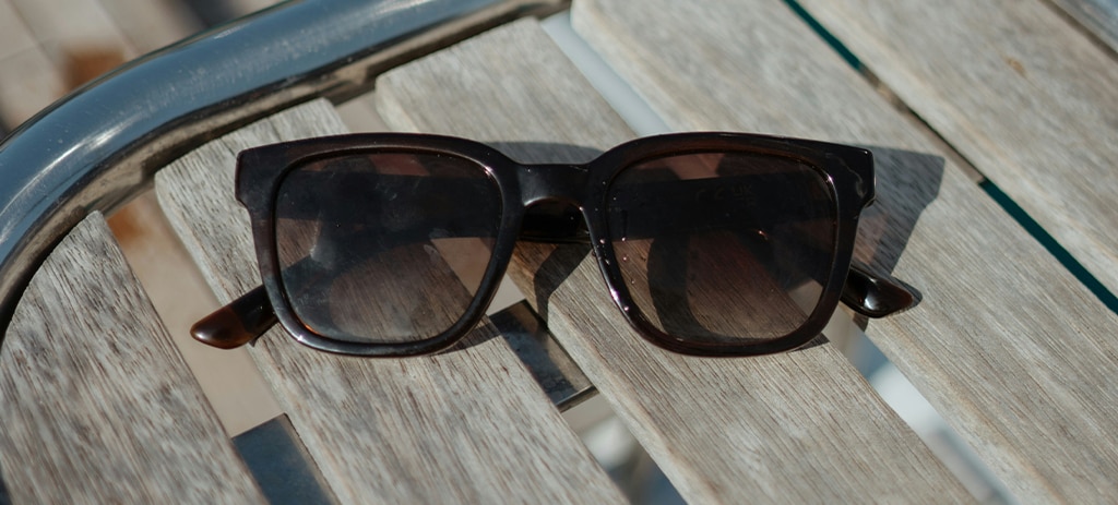 A close-up of black square acetate sunglasses with brown lenses, resting on a slatted wooden surface.