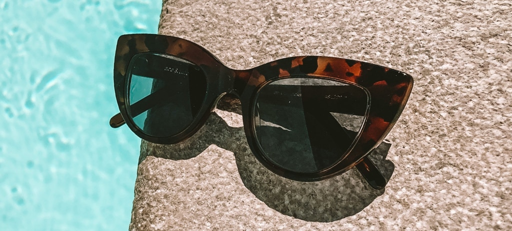 A pair of brown Havana cat-eye designer sunglasses with grey lenses rest stylishly at the edge of a pool.