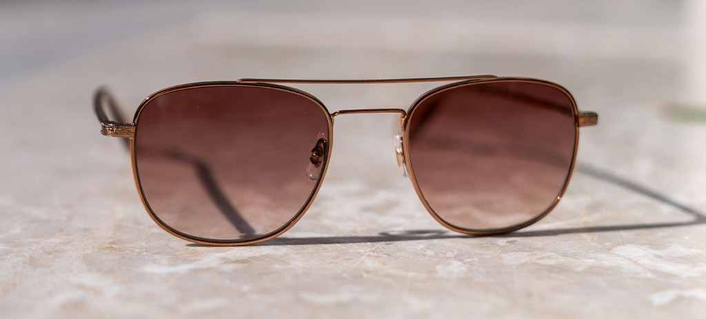 A pair of pilot-style sunglasses resting open on a marble-like surface, facing away from the camera. The sunglasses are a bronze metallic colour with ruby-coloured lenses. 