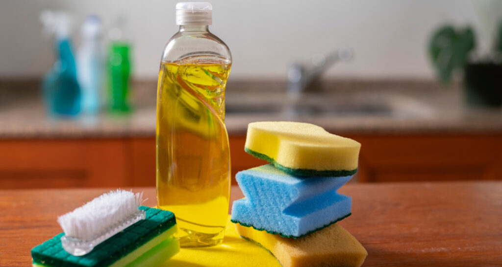 A close-up of a clear bottle with yellow washing-up liquid in, flanked by a stack of three sponges on one side and a sponge with a brush on the other. In the softly blurred, well-lit indoor background, a kitchen sink is faintly visible.