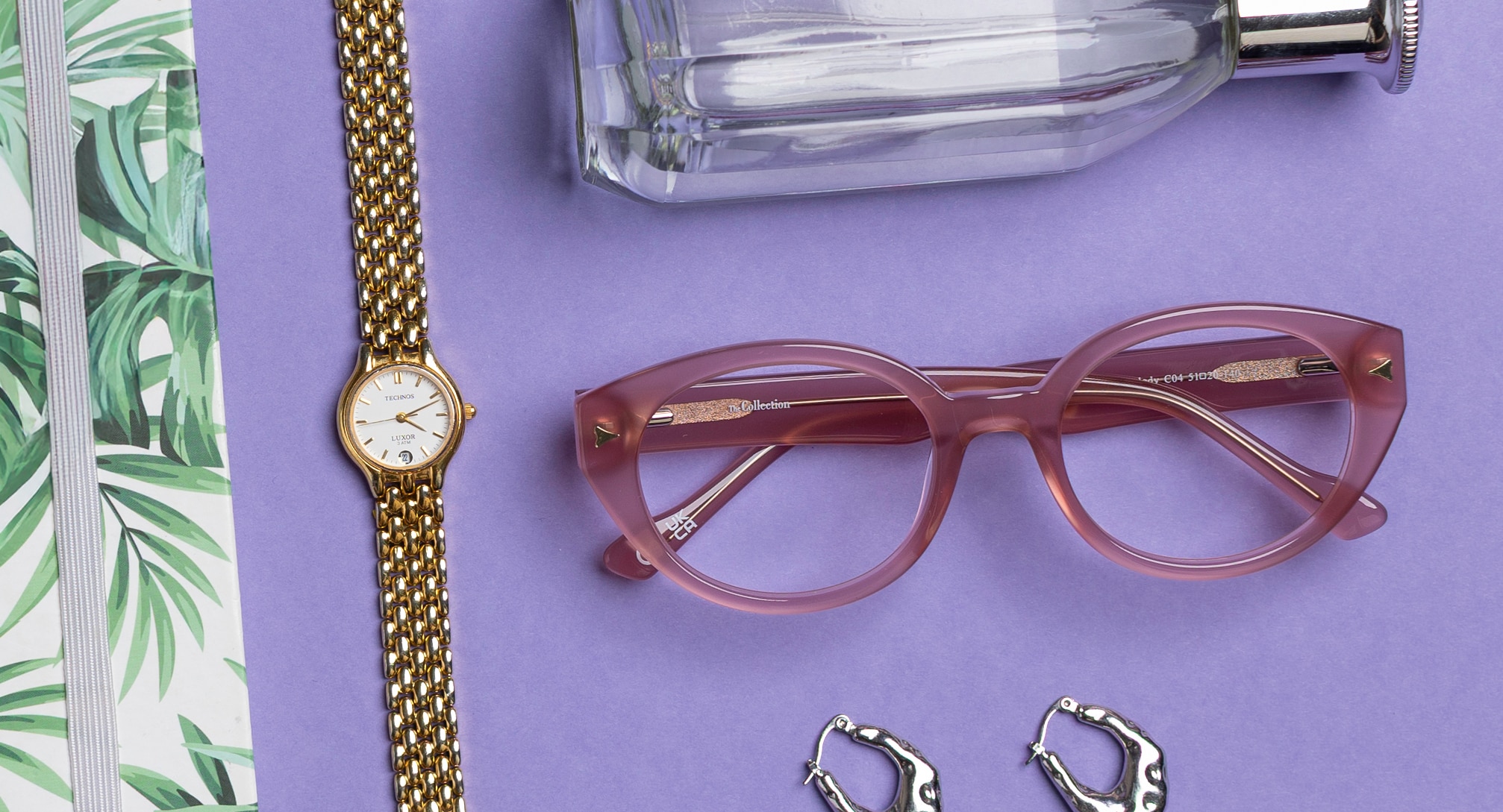 At the centre of a lavender background rests a pair of milky pink oval frames, accompanied by a gold metal wristwatch. Around the edges, glimpses of essentials appear—a white notebook with a green leaf print, gold hoop earrings and a clear perfume bottle.