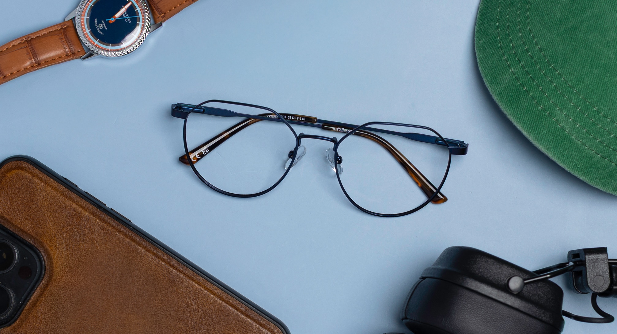 A pair of blue metal phantos-shaped frames sit at the centre of a light blue background, surrounded by several essential items which can just be seen at the edges, including a brown leather wrist watch, mobile phone in a brown leather case, black over-ear headphones and a green baseball cap. 