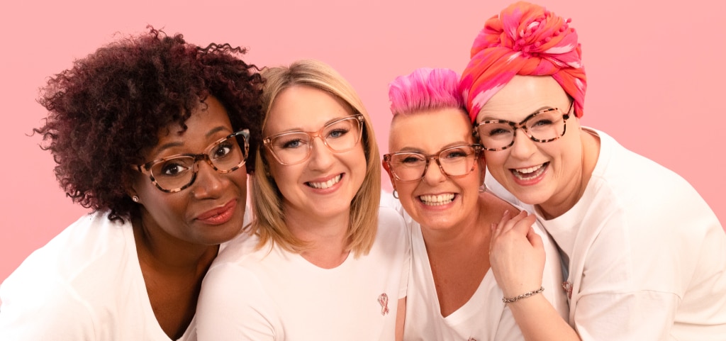 A close-up of four women in plain white t-shirts, each wearing the Pink Ribbon badge, smiling warmly against a pastel pink backdrop. On the far right, a woman with dark curly hair and red lipstick wears pink Havana cat-eye glasses. Beside her, a blonde woman with straight, medium-length hair sports transparent pink frames. Next, a woman with a bright pink quiff wears brown striped cat-eyes. On the far left, a woman with a colourful pink headscarf completes the group with square pink Havana glasses.