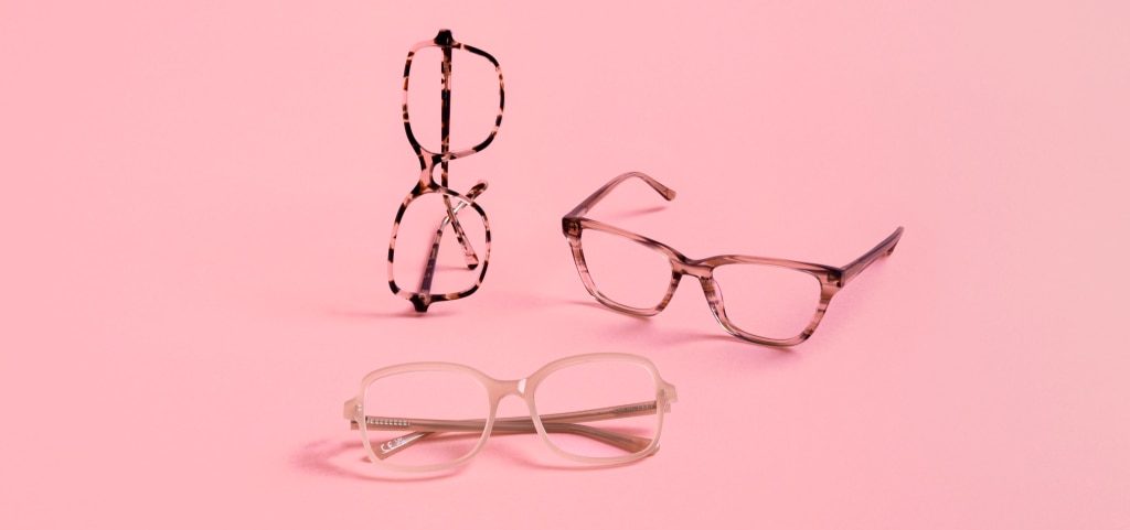 Three Pink Ribbon acetate glasses are displayed against a pastel pink background. One pair, a pink Havana square frame, stands upright on its side. Beside it rests a pink and brown striped cat-eye frame, while in front, a nude square frame sits neatly folded.