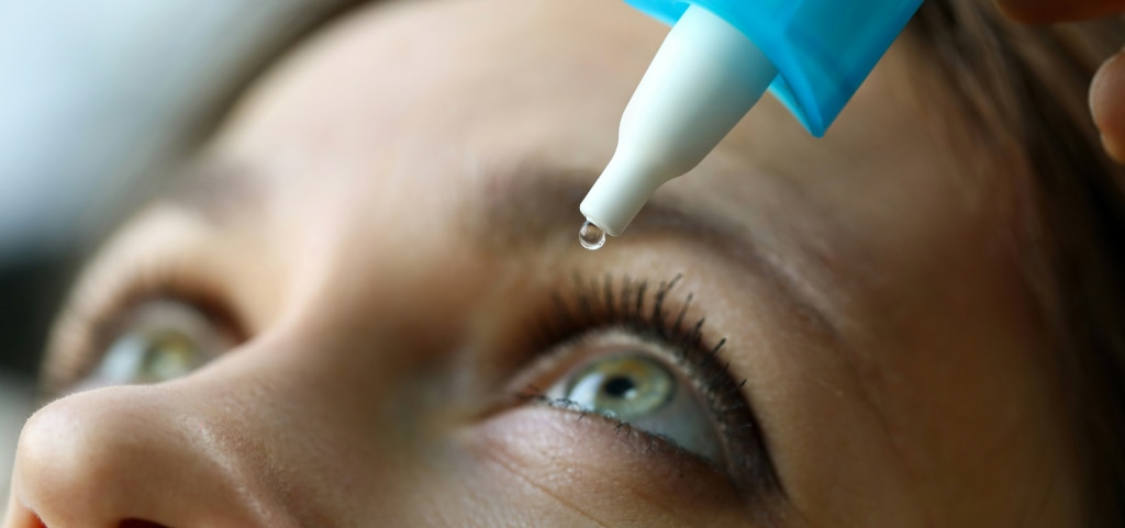 A close-up of a woman applying eye drops to her left eye. She has light brown hair and striking green eyes.
