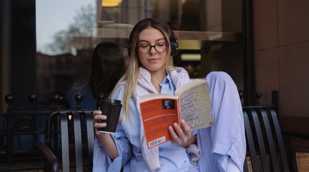 A young woman sits outdoors on a black metal bench, wearing a white and blue striped co-ord, round acetate glasses and black over-ear headphones. She holds an open book in her left hand and a coffee cup in the other, enjoying the bright setting.