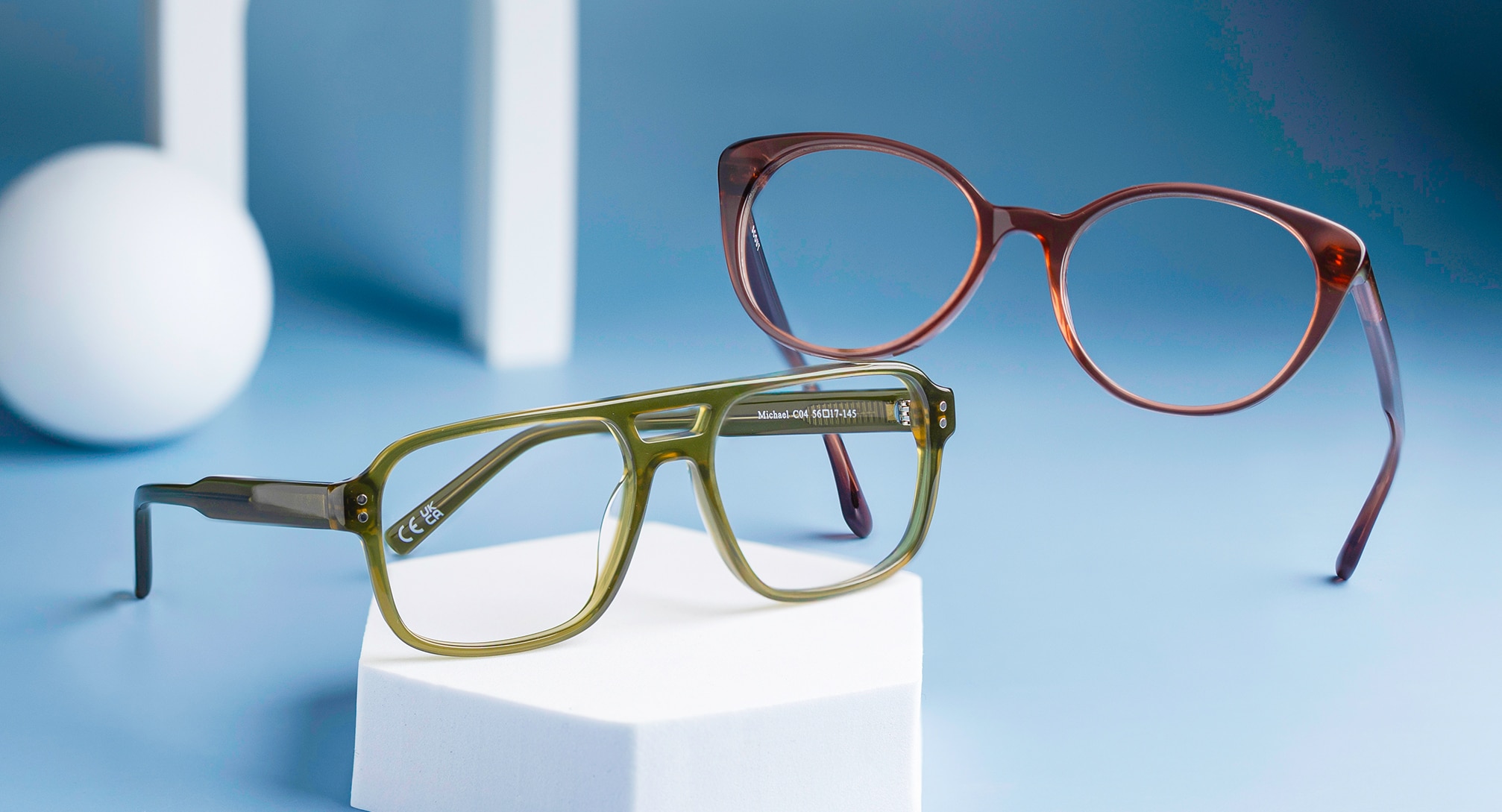 Two colourful bio-acetate frames from the Scout range are displayed on a 3D hexagon against a light blue background, with a 3D ball and ridge visible behind. One features a green pilot design with metal rivets, while the other, a pair of crystal purple cat-eye frames, rest elegantly on top.