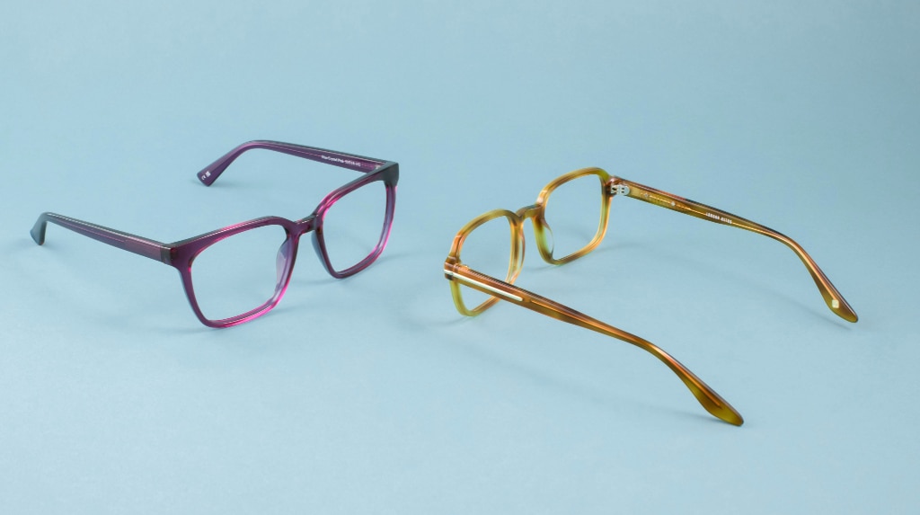 Two pairs of glasses rest open on a light blue background, facing each other. One is a square purple acetate frame, the other a honey-toned rectangular acetate design with metal temple accents.