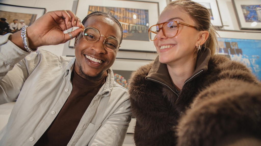 In a bright indoor setting, two young people sit together, smiling. Behind them, framed pictures decorate the wall. The young man, with dark dreadlocks swept back, wears gold metal geometric glasses, a brown T-shirt layered under a beige shirt and a chunky silver chain bracelet. Beside him, the young woman wears orange acetate pilot glasses and a brown fur coat, extending her arm toward the camera as if snapping a selfie.