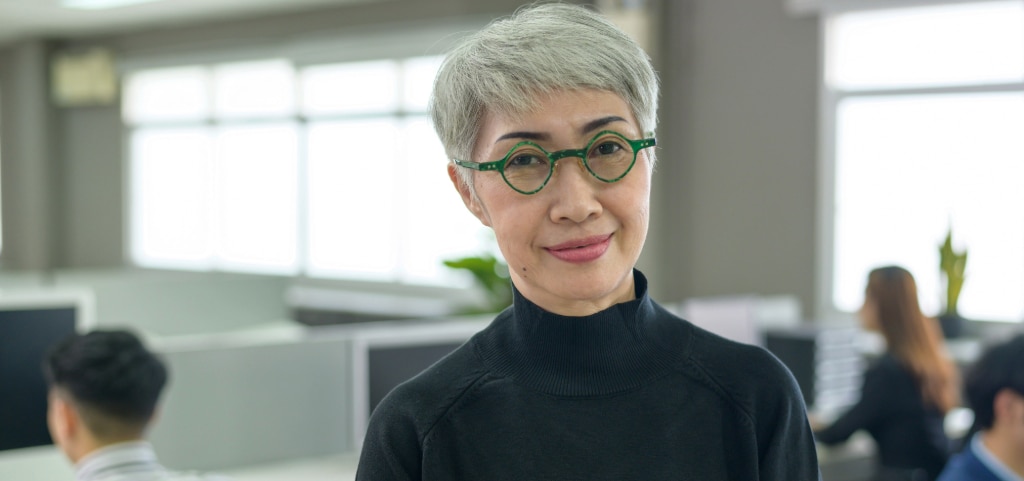 A close-up of a middle-aged Asian woman in a bright office setting. She has short grey hair and is wearing round green acetate glasses with a black polo-neck jumper.