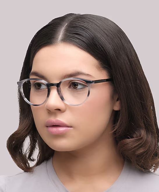 A portrait of a young woman with short, dark hair against a white background. She’s wearing small grey phantos-shaped glasses and a grey t-shirt, gazing off to the right.