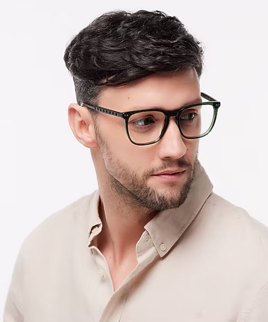 A portrait of a young man with short, dark undercut hair against a white background. He’s wearing large, square green glasses and a beige shirt, gazing off to the far right.