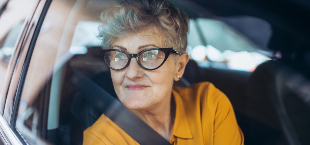 A close-up of a woman with short, well-styled grey hair wearing black cat-eye glasses and a mustard yellow blouse, looking out the car window with her seatbelt fastened.