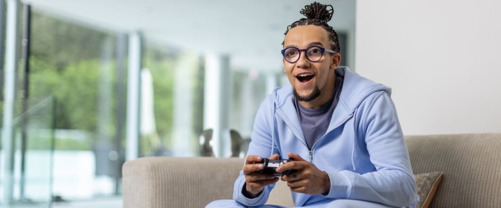 A young man with dreadlocks tied in a bun sits on a beige sofa in a bright, modern room. He wears bright blue round acetate glasses, a light blue tracksuit, and a navy t-shirt. His mouth is open in surprise as he grips a gaming controller.