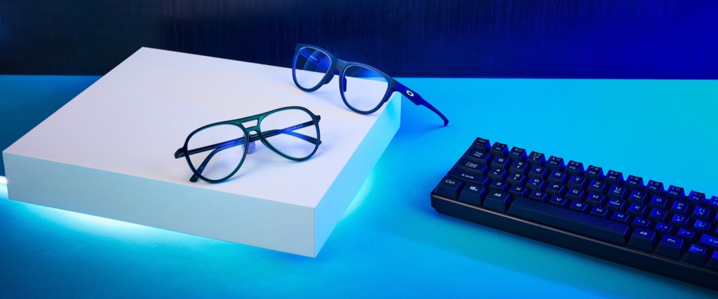 Two pairs of glasses rest on a white box illuminated by a soft blue LED light from beneath. One pair, made of blue plastic with a rounded square shape and the Oakley symbol on the temples, lies open and balanced on the edge. The other, a blue plastic aviator style, is folded neatly in the centre. A black keyboard sits to one side.