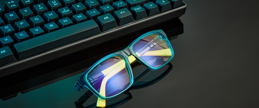 A pair of transparent green and yellow bio-nylon glasses, with blue light lenses, lies folded on a black surface, with a black keyboard visible just above them.