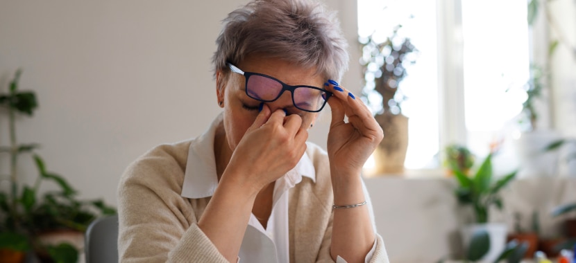 A close-up of a woman with short grey hair holding her glasses out of the way with one hand while rubbing her eyes  with the other. She’s wearing a white blouse and a beige cardigan in a bright indoor setting.