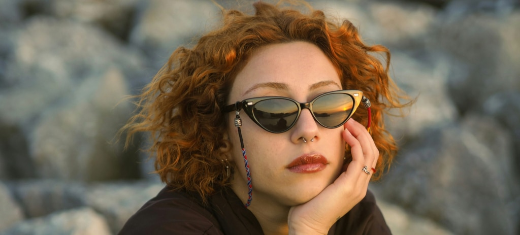 A young woman with short, curly red hair rests her head on one hand while sitting in a bright outdoor setting, wearing a brown jacket and black Ray-Ban cat-eye sunglasses with a multi-colour strap.