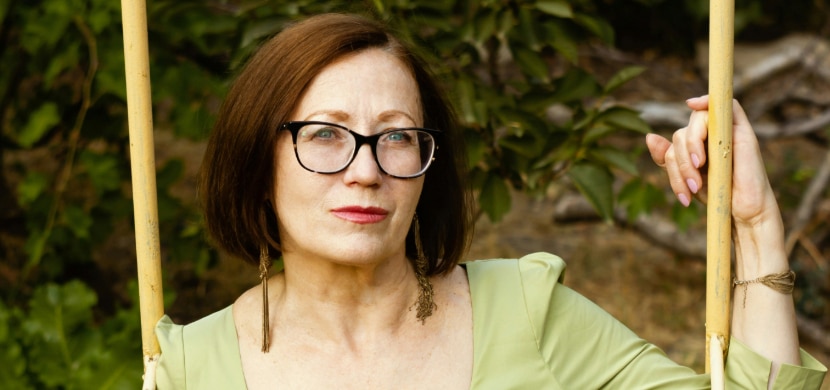 A close-up of a confident middle-aged woman with a tidy brown bob, sitting on a swing in a bright outdoor setting. She’s wearing rectangular tortoiseshell acetate glasses and a light green dress.