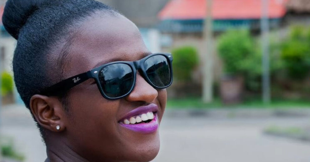 Close-up of a young woman with black hair in a bun, wearing black Ray-Ban Wayfarers in a bright outdoor setting. She smiles brightly with pink lipstick.