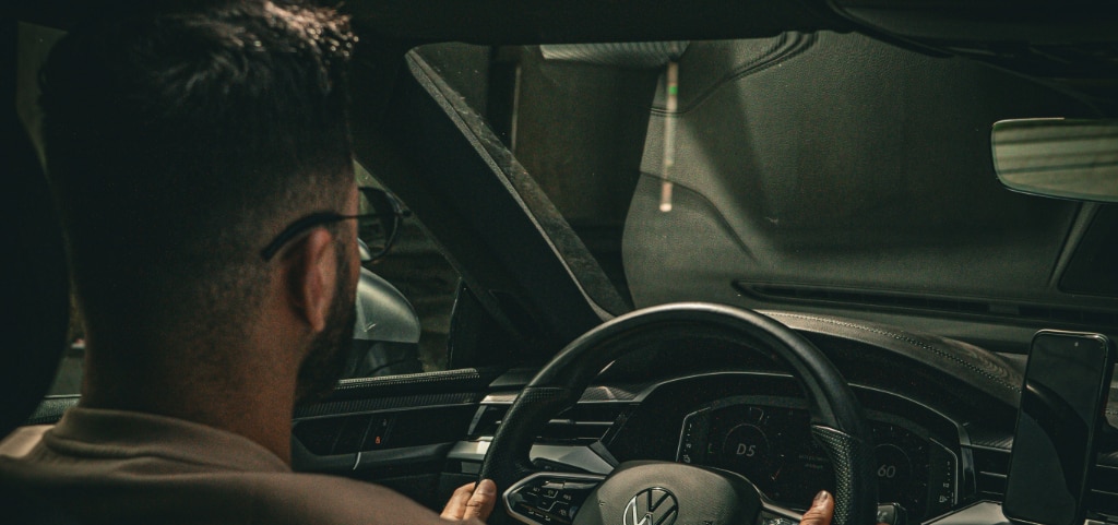 A close-up of the back of a young driver’s head as he drives at night. He has short undercut hair, a beard, and wears round metal glasses.