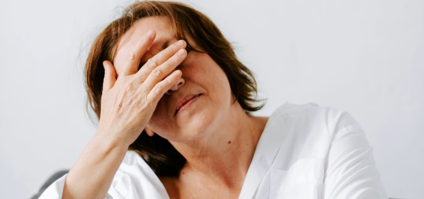 A close-up of a middle-aged woman with medium-length brown hair, wearing a loose white blouse, holding one hand over her face as though fatigued.