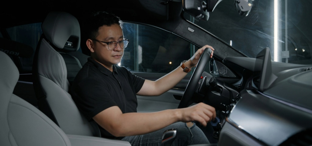 A passenger-view shot of a young Asian man driving a car in low light. He has short dark hair and wears square black metal-rimmed glasses, a black polo shirt, and grey ripped jeans. One hand rests on the steering wheel while he adjusts the dashboard controls with the other.