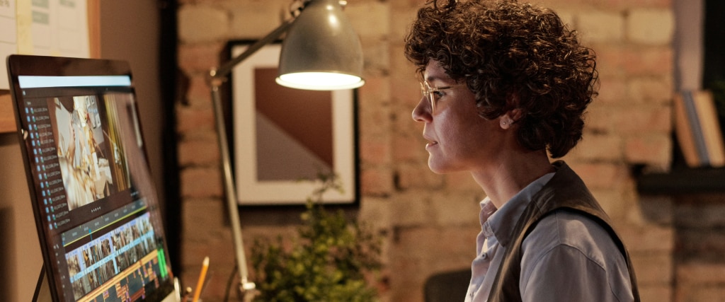 A side close-up of a young lady with short brown curly hair in a bright indoor setting. She is sitting looking at a Mac computer screen with a lamp beside her. The lady is wearing round acetate glasses and a pale shirt and waistcoat.